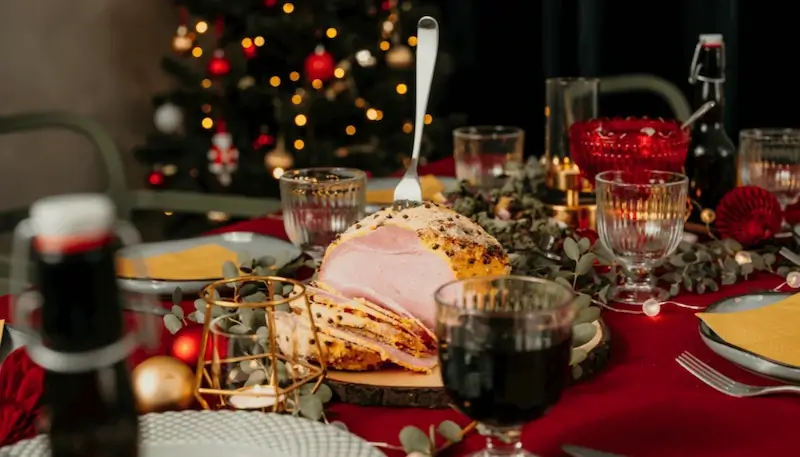 A roast ham on a Christmas table with a Christmas tree at the back and wine glasses in the forefront