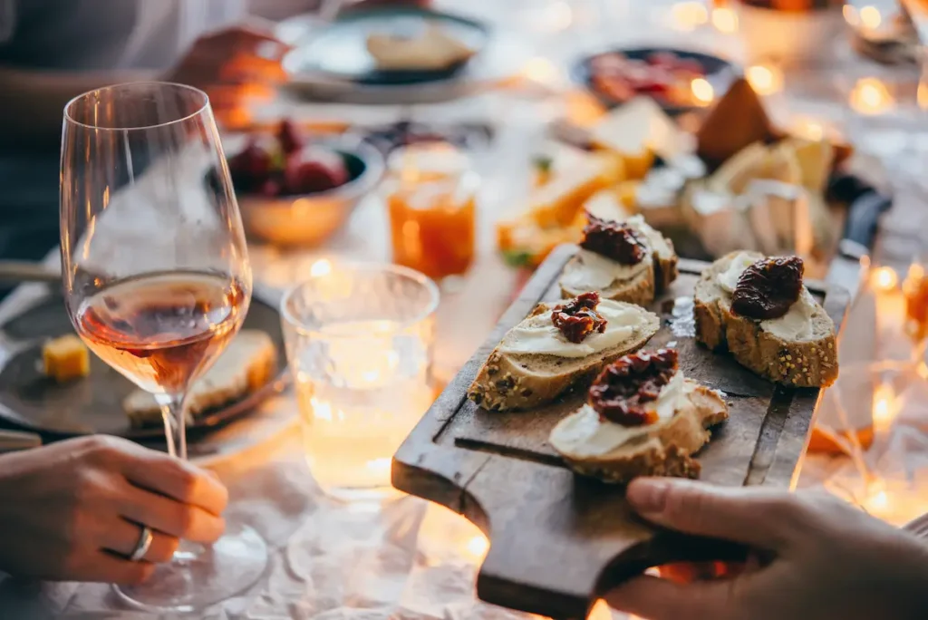 A table decorated with lights and candles. Close up of a wine glass with white wine held by a hand. Another person is passing a wooden board around the table, with bread and cheese.