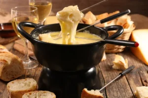 A piece of bread being dipped in a cheese fondue pot, surrounded with pieces of bread alll over the wooden table