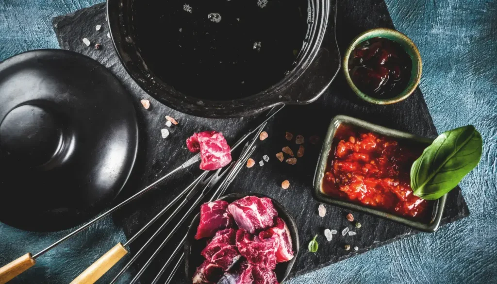 A pot of hot oil for meat fondue with several pieces of meat on fondue skewers and in plates. The pot is set up on a table.