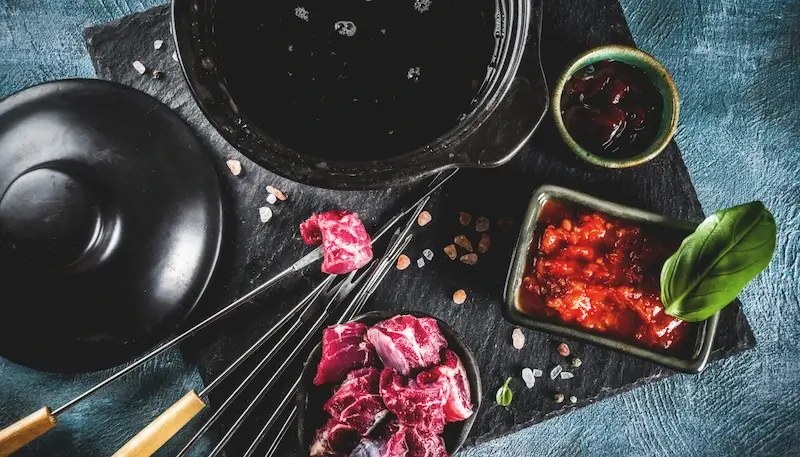 A pot of hot oil for meat fondue with several pieces of meat on fondue skewers and in plates. The pot is set up on a table.