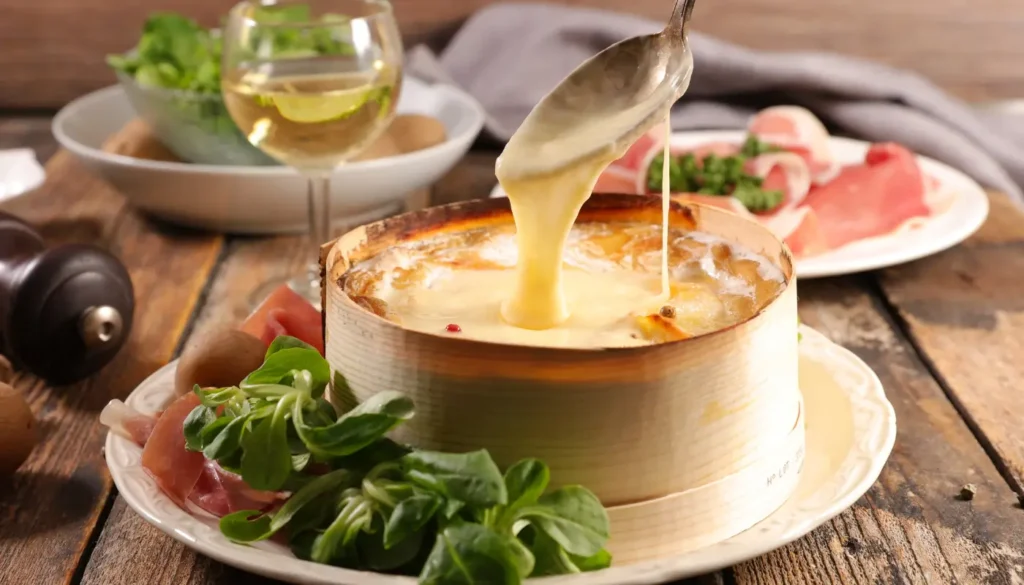 A table with a baked Mont d'Or, charcuterie and wine. A spoon is being dipped in the cheese to show how it is melted.