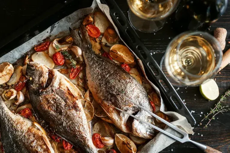 Baked fish in a pan with roasted tomatoes. The pan is on the table, with some white wine glasses next to it.