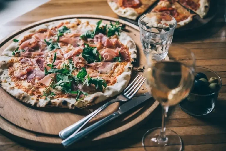 A ham and cheese pizza on a table with a glass of white wine next to the pizza board