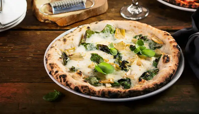 A white-based pizza, with some spinach and artichokes, in a plate on a table