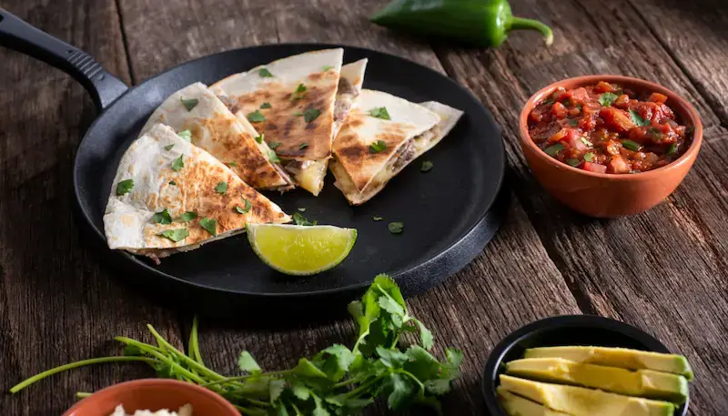 A table with quesadillas on a plate and some bowls with chillies and other condiments