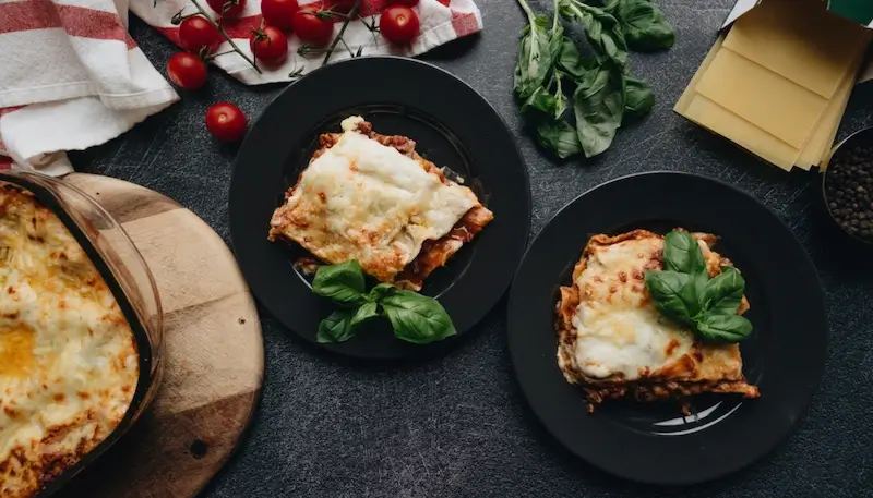 A table with a lasagna dish and two plates with a slice of lasagna. Some ingredients are placed on the table - tomato, basil, lasagna sheets, to illustrate.